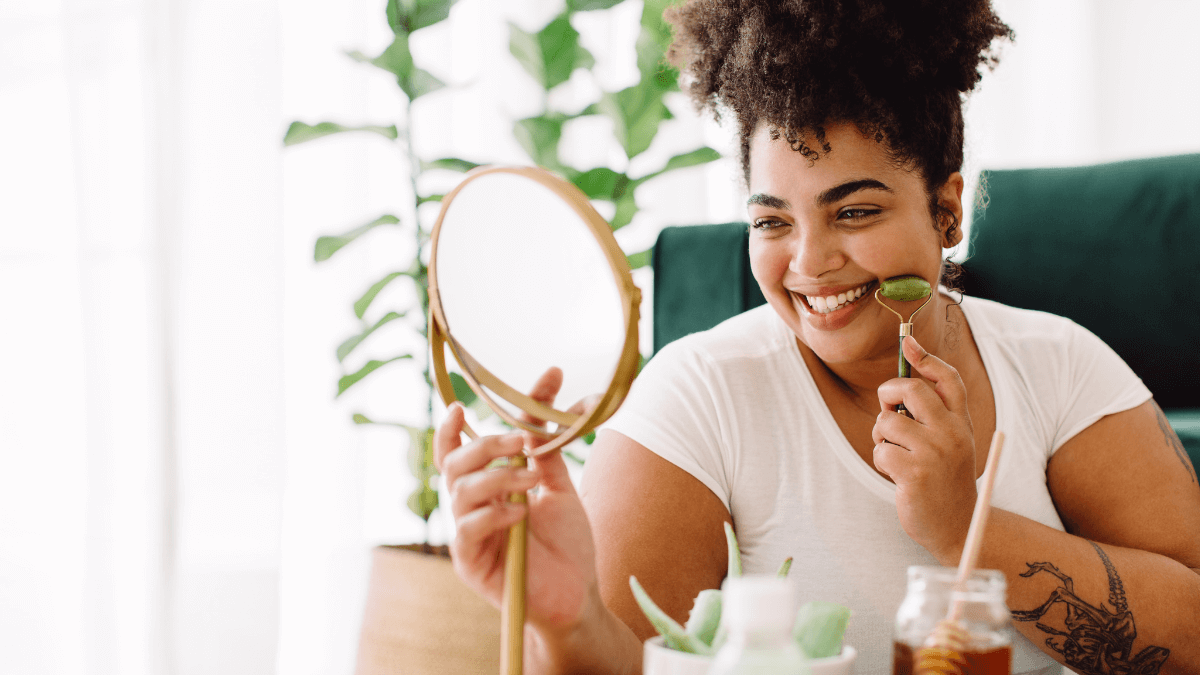 A young woman is smiling and looking in the mirror while practicing self-care for a glowing complexion, embracing the joy of solo living.